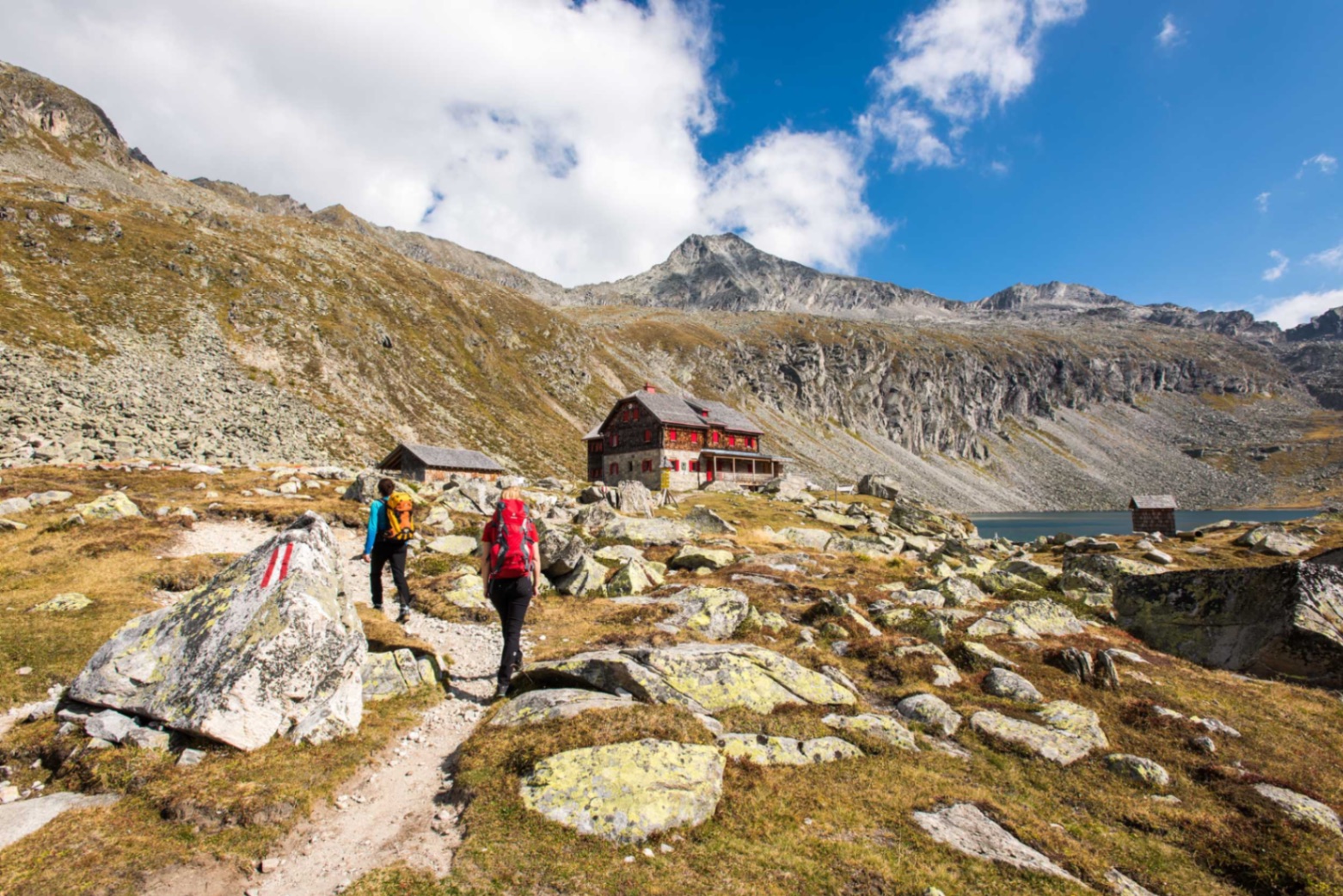 Wanderer auf dem Weg zu einer Berghütte vor See und Bergpanorama